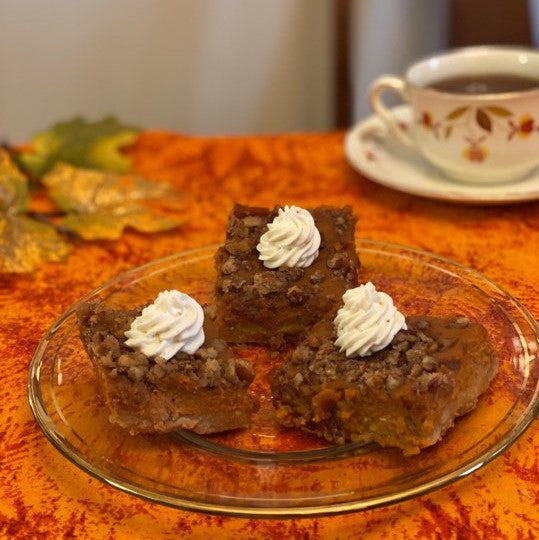 Image of 3 pumpkin pie squares with whipped cream piped on top. Orange and red speckled table cloth with leaves  and a cup of tea in the background.